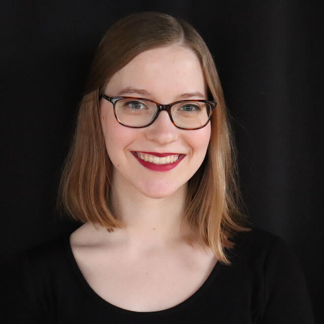 Headshot of Sabine. She is wearing brown glasses and red lipstick. Her hair is shoulder-length, and she is smiling.